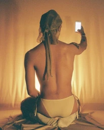 Shirtless man sitting on a bed with a head covering holding up a lit smartphone to take a photo, shot by Clifford Prince King.
