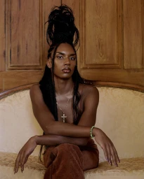 Portrait of a Black woman with tall braided updo wearing a cross necklace and seated on a couch by photographer Clifford Prince King