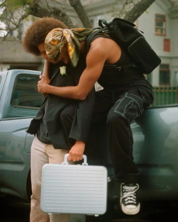 Two black men embracing outdoors with one holding a silver briefcase near a dark pickup truck, photographed by Clifford Prince King