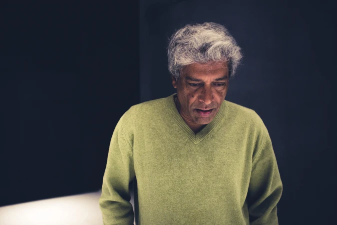Portrait of a mature man with gray hair, wearing a light green sweater, standing in front of a dark background in a studio setting.
