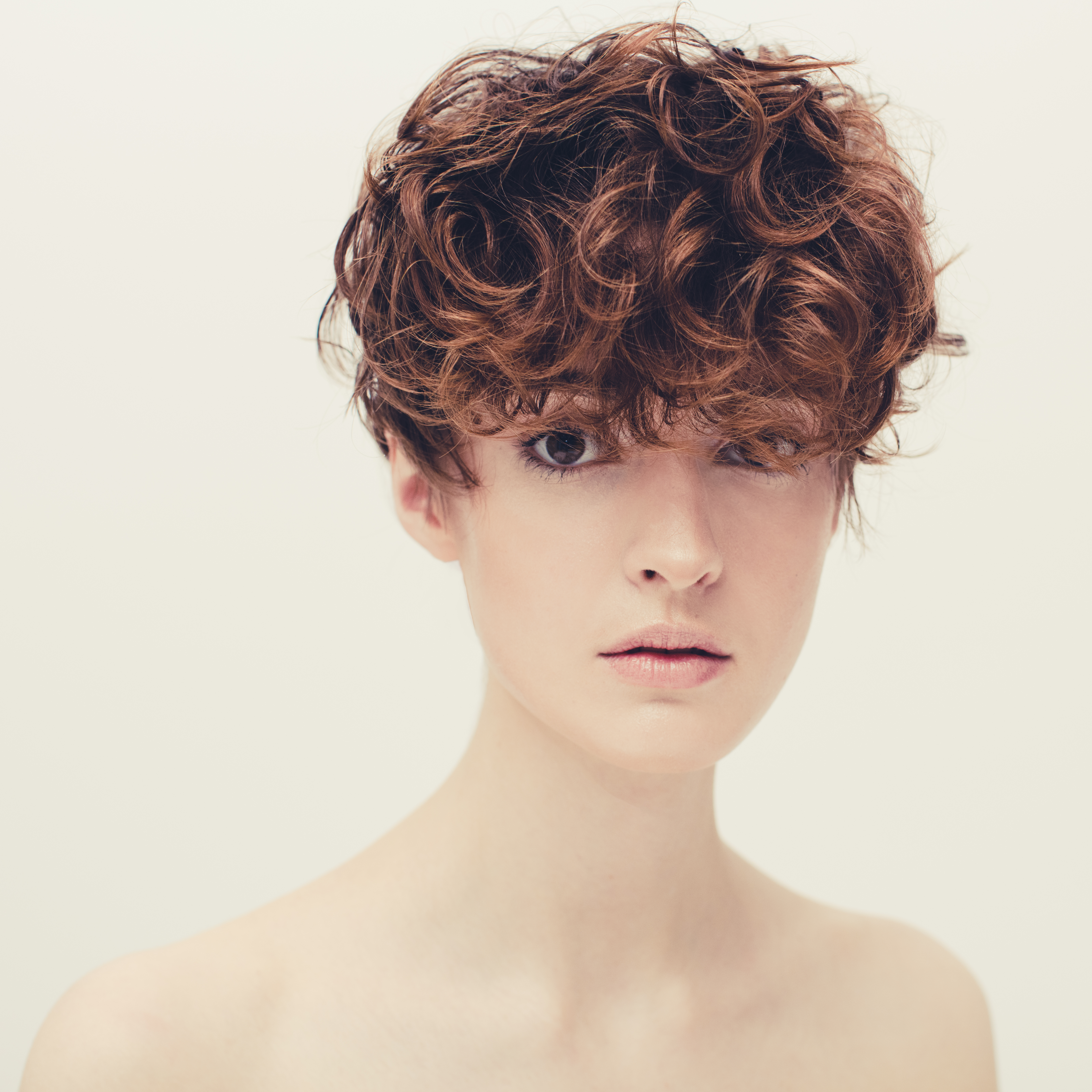 Portrait of a fair-skinned woman with curly auburn hair and bare shoulders, looking directly at the camera against a light backdrop.