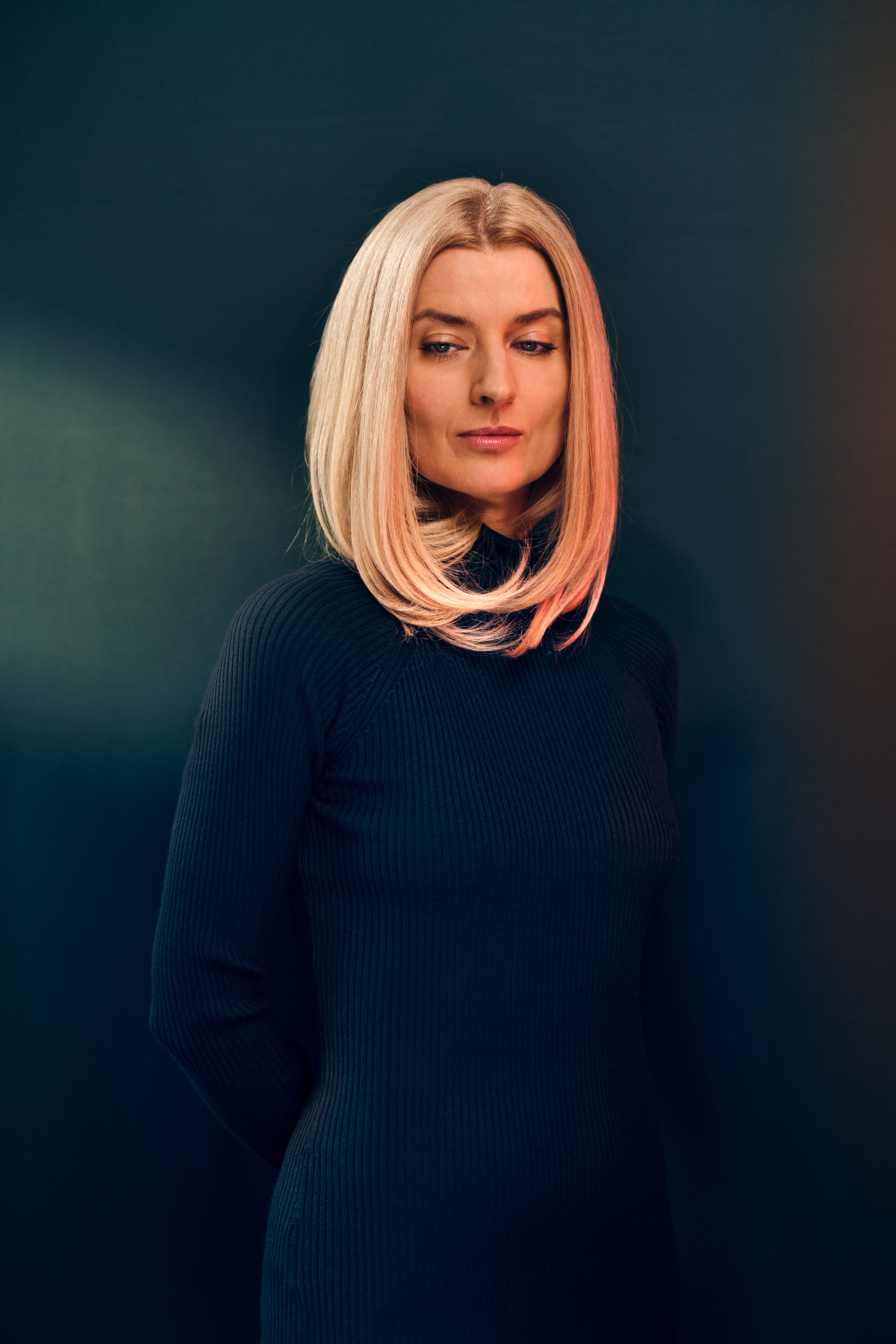 Portrait of a blonde woman in a black dress against a dark teal backdrop, illuminated with a warm light. Mid shot, eye-level angle.