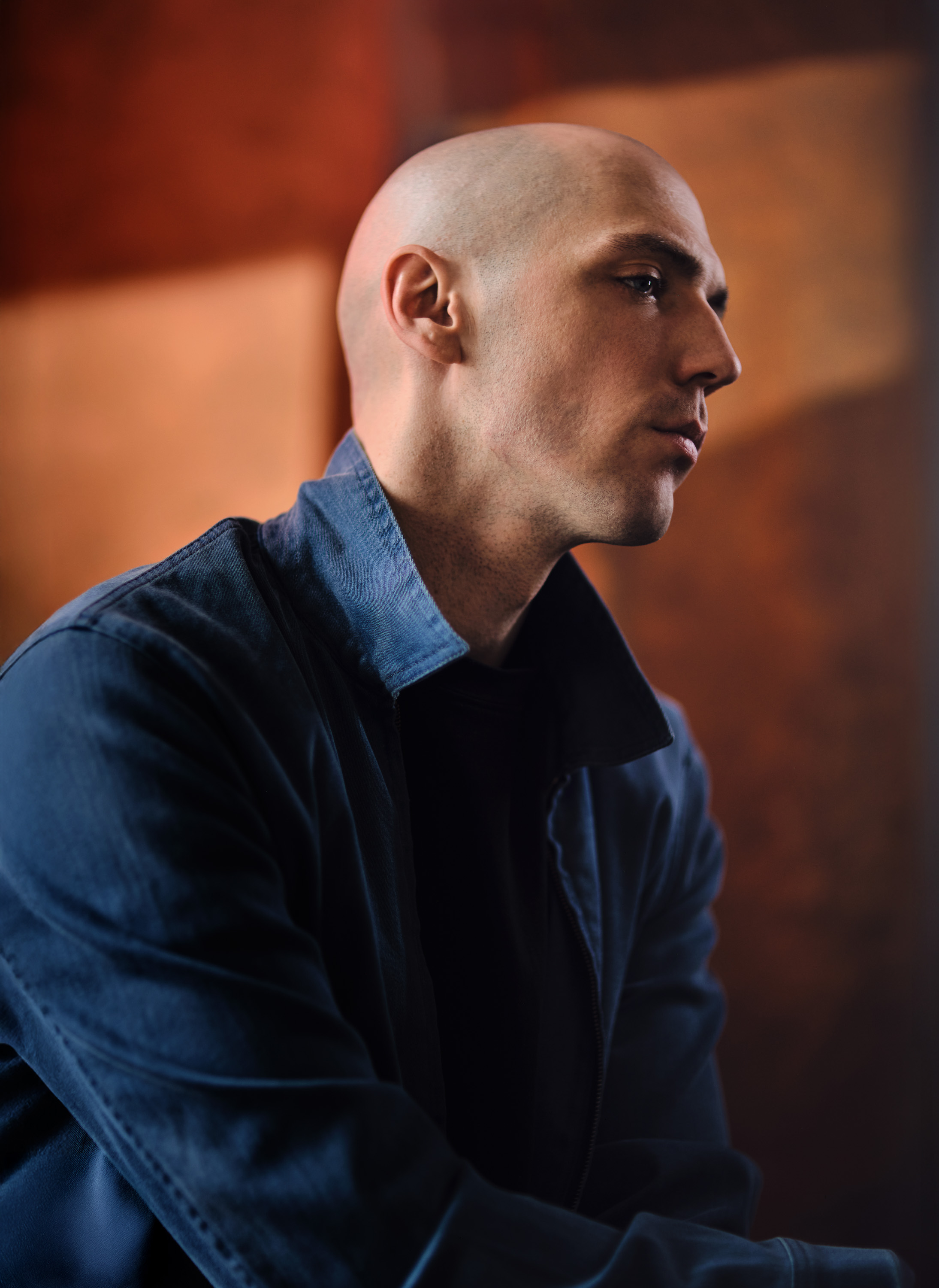 Portrait of a bald adult male with a blue collared shirt, gazing off-camera. Dark background with brown and orange hues, subtle gradient lighting.