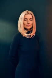 Portrait of a blonde woman wearing a black turtleneck dress against a dark wall with subtle pink and blue lighting.