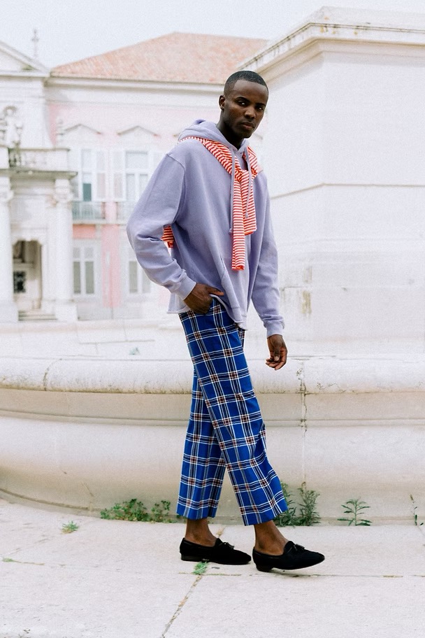 Full body shot of a Black male model in a lilac hoodie and plaid pants, posing on a concrete structure with a building behind.