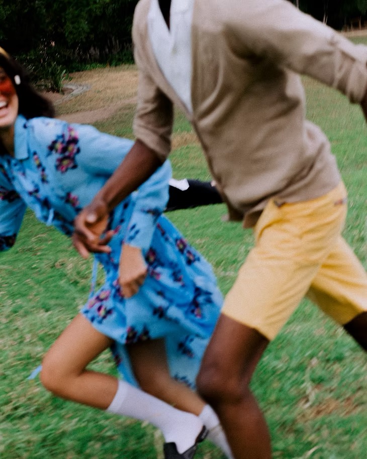 Two adults joyfully run on a green lawn, one in floral dress and other in yellow shorts and cardigan, with soft, natural lighting.