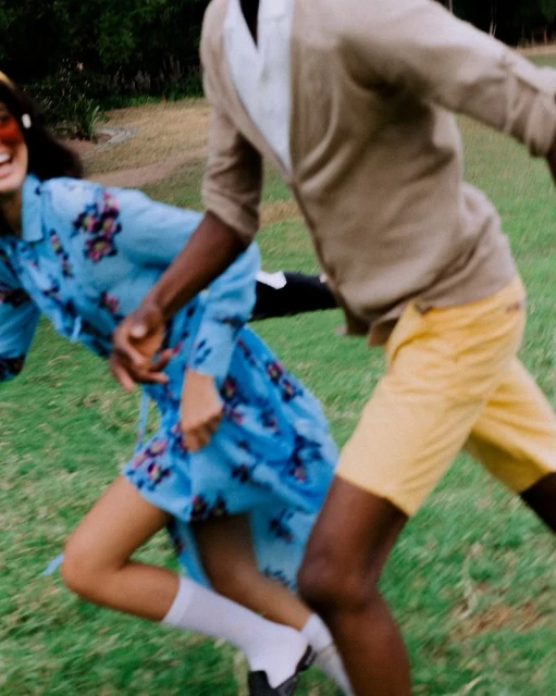 Two adults joyfully run on a green lawn, one in floral dress and other in yellow shorts and cardigan, with soft, natural lighting.