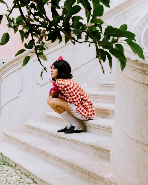 Young woman in patterned coat sits on stone steps outdoors with greenery, looking left in a fashion style photo.