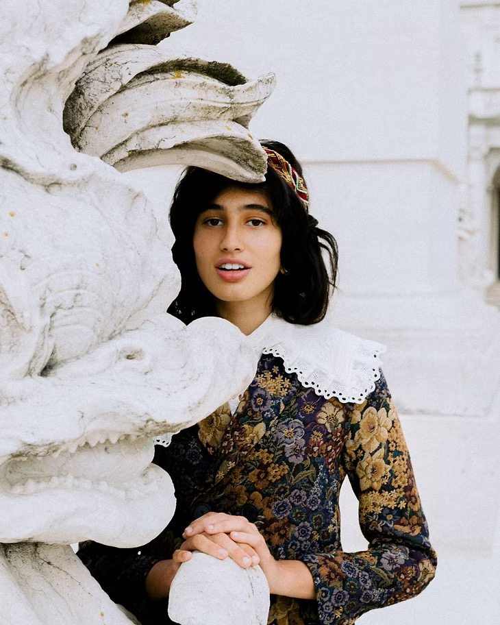 Portrait of a woman with dark hair wearing a floral dress and lace collar standing near a stone sculpture outdoors in daylight.