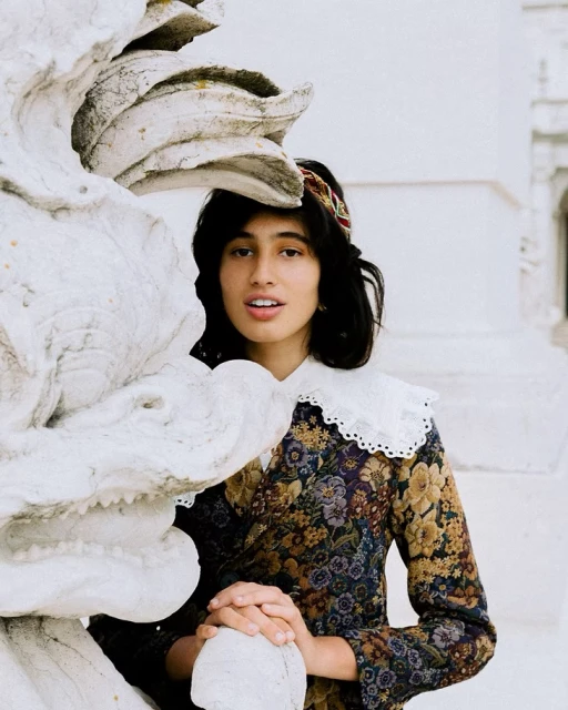 Portrait of a woman with dark hair wearing a floral dress and lace collar standing near a stone sculpture outdoors in daylight.