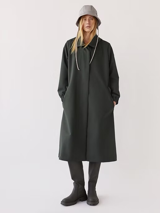 A full-length studio shot of a woman in a dark green coat and bucket hat, standing against a white background, showcasing contemporary fashion.
