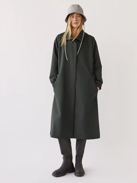 A full-length studio shot of a woman in a dark green coat and bucket hat, standing against a white background, showcasing contemporary fashion.