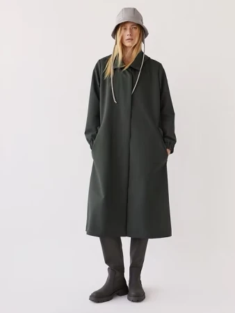 A full-length studio shot of a woman in a dark green coat and bucket hat, standing against a white background, showcasing contemporary fashion.