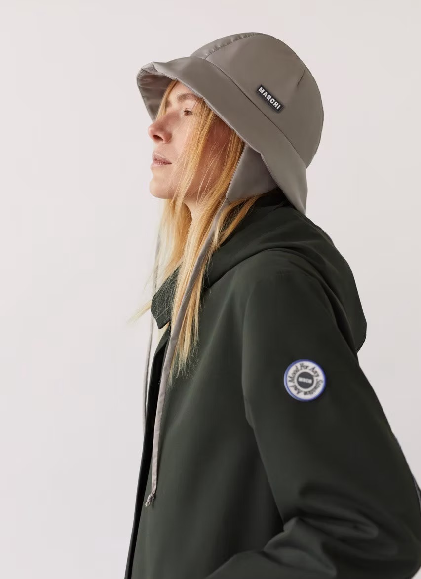Woman in profile wearing grey bucket hat with ear flaps and a dark green hooded jacket against a plain white background in a studio shot.