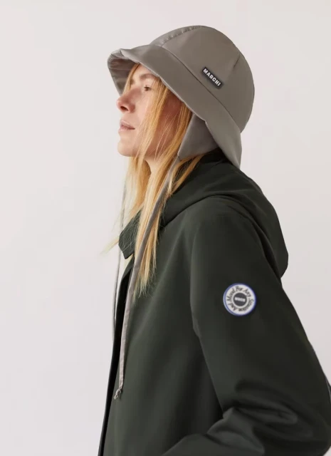 Woman in profile wearing grey bucket hat with ear flaps and a dark green hooded jacket against a plain white background in a studio shot.