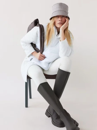 Model seated on a chair wearing a white quilted jacket, grey bucket hat, ribbed tights, and tall dark boots.