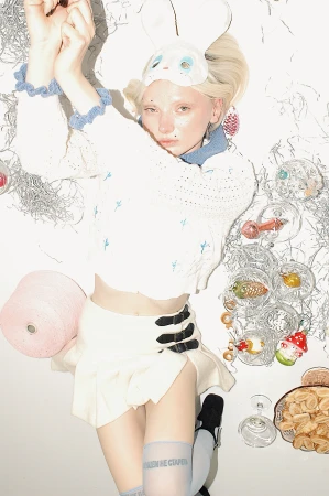 Fair-skinned person lying on a white surface wearing a bunny mask, white knit sweater, mini skirt, and knee-high socks, surrounded by props.