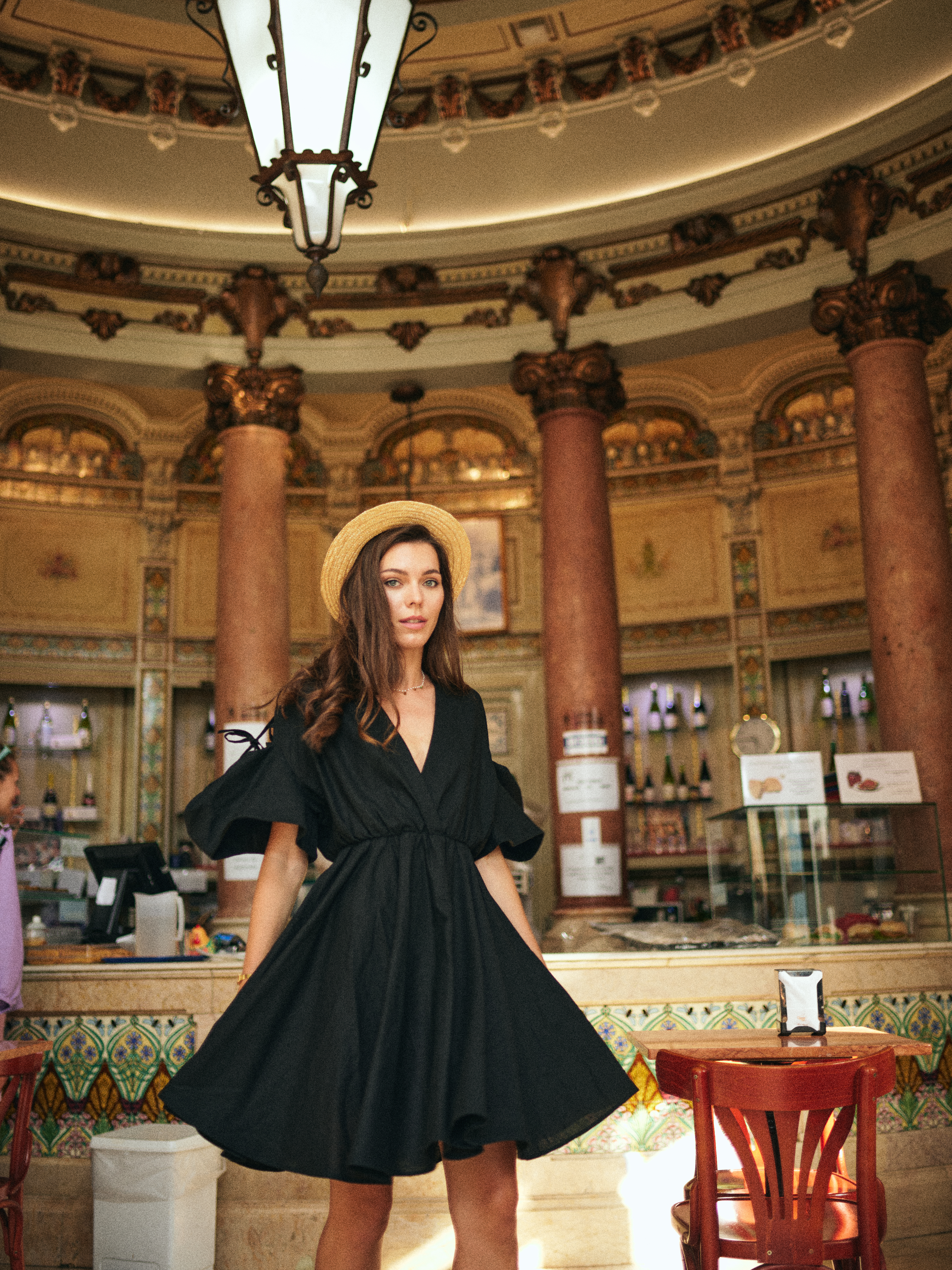 A woman wearing a black dress and straw hat poses inside an ornate, high-ceilinged building with columns, tables, and a counter with bottles.
