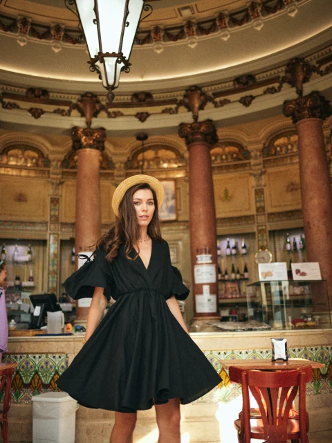 A woman wearing a black dress and straw hat poses inside an ornate, high-ceilinged building with columns, tables, and a counter with bottles.