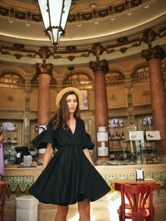A woman wearing a black dress and straw hat poses inside an ornate, high-ceilinged building with columns, tables, and a counter with bottles.
