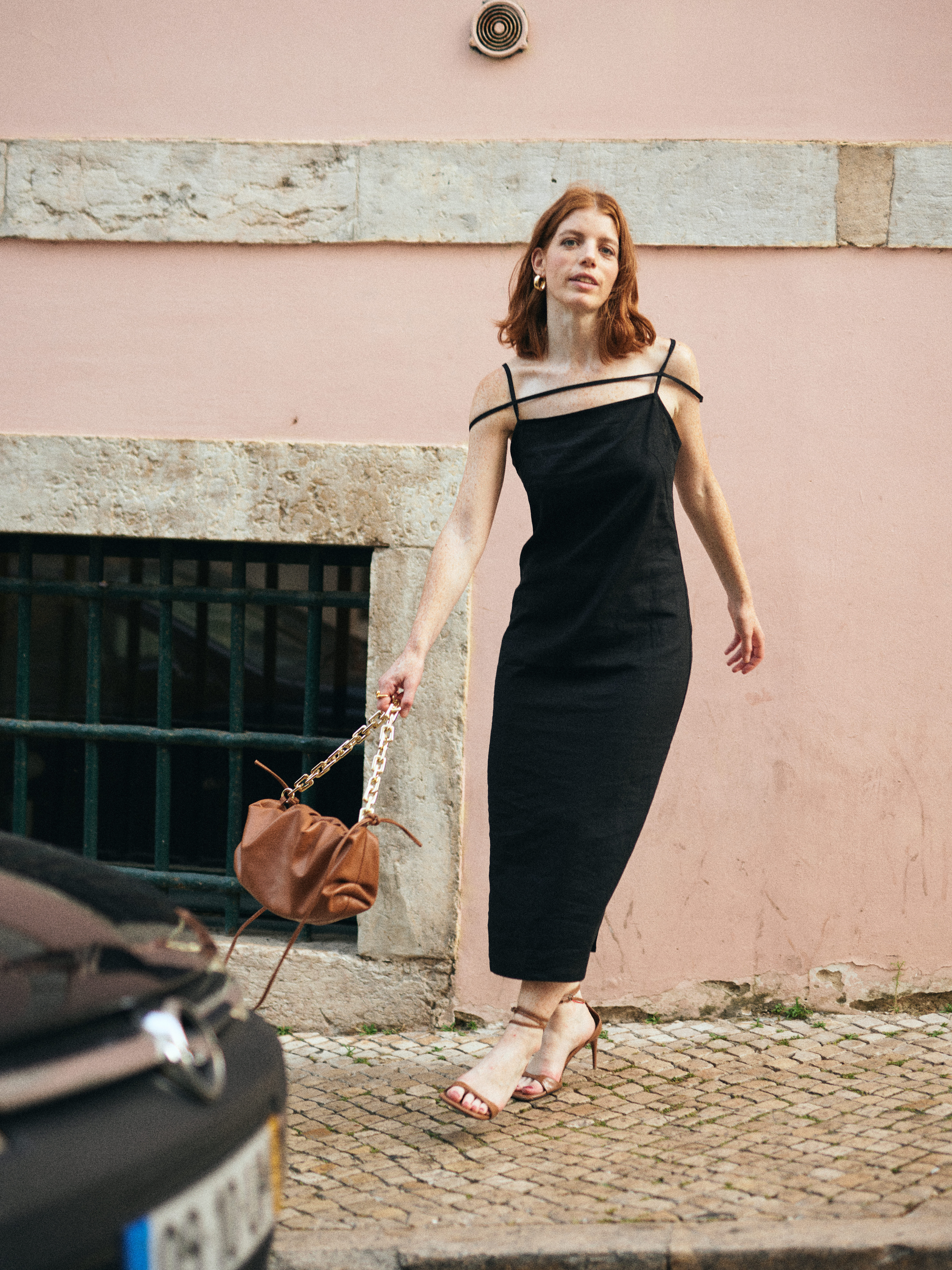 A red-haired woman in a black dress and heels stands outside next to a car, carrying a brown purse with a chain strap.