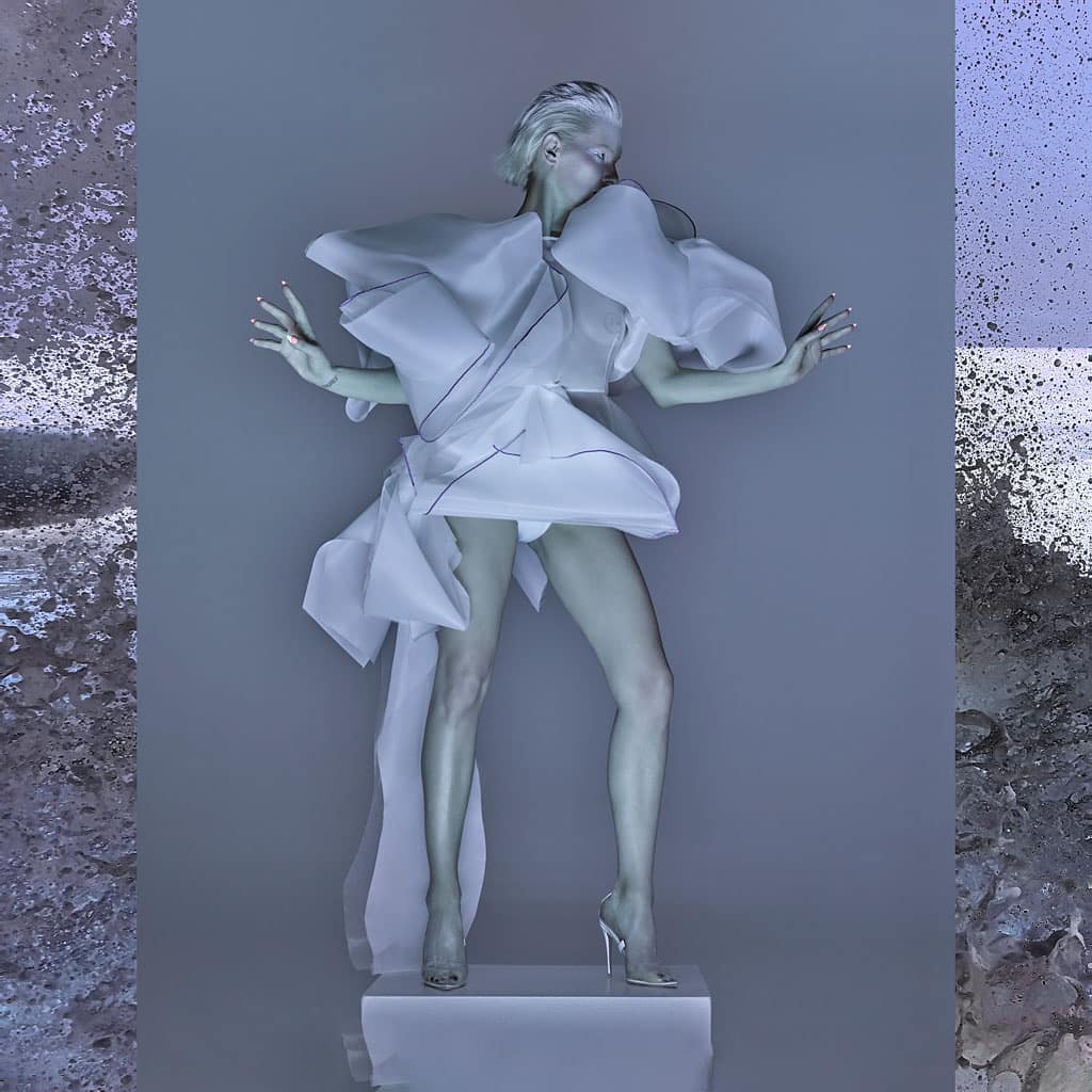 Adult female model poses in a white sculptural dress on a white cube against a grey background with textured accents on either side.