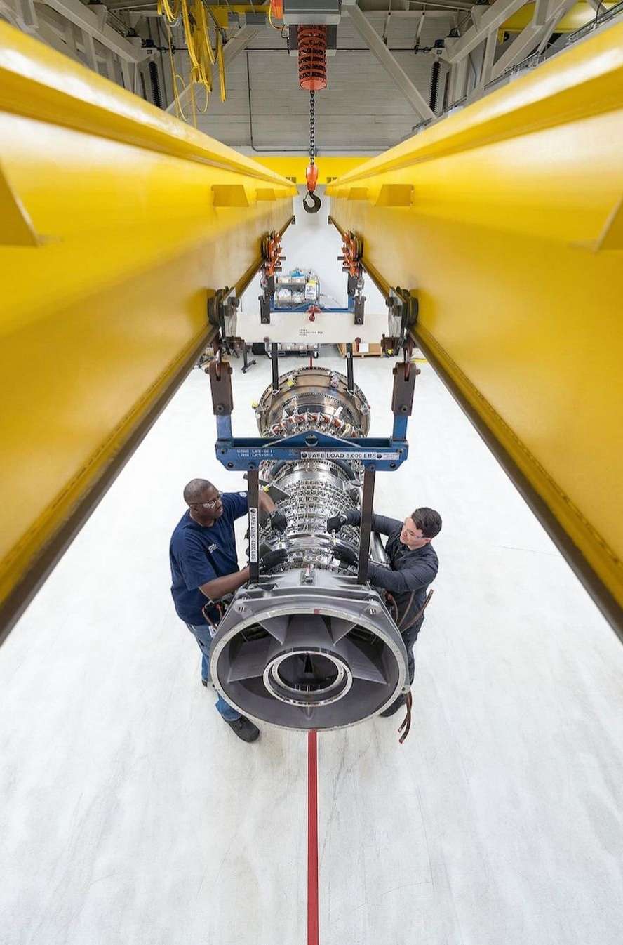 Two engineers inspect a large, silver jet engine suspended by a yellow crane in an industrial setting, showcasing aerospace maintenance.