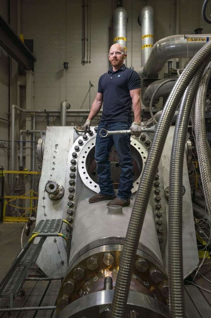 Man standing on industrial machinery, holding a wrench. He wears a dark shirt and jeans in an industrial setting with metal piping.