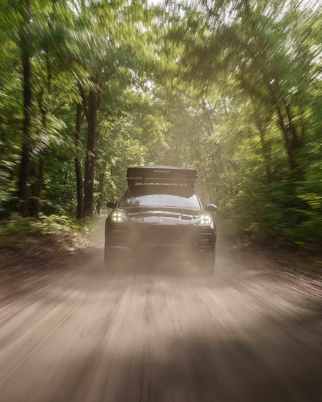Dark grey SUV with rooftop cargo carrier driving on a dirt road through a bright green forest with motion blur to imply speed.
