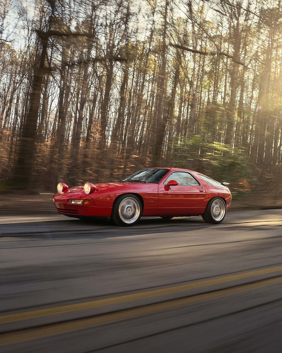 Red Porsche 928 driving on a rural road flanked by bare trees, captured in motion with blurred wheels and sunlit background.