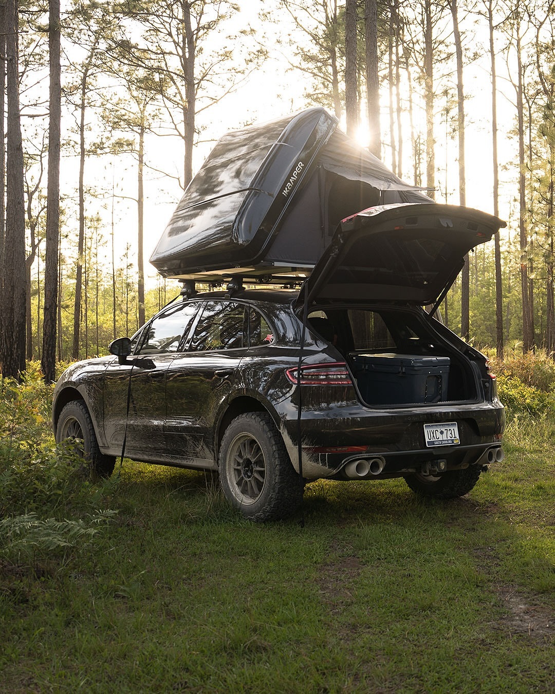 Black SUV with roof tent parked on grass in forest. Car has open trunk and off-road tires, lit by sun in background. Camping concept.