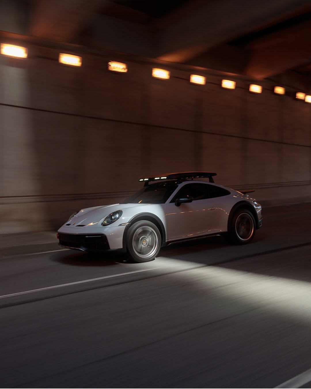 White Porsche 911 Dakar driving through a tunnel, headlight beams visible, emphasizing motion and the car's design, ambient tunnel lighting.