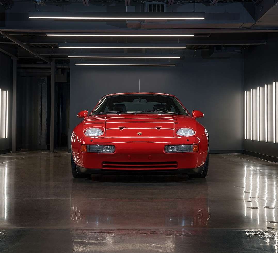Front-facing view of a shiny, red 1980s Porsche 928 parked in a modern-looking indoor garage with glossy floors and bright ceiling lights.