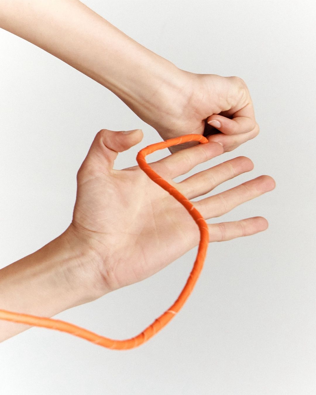 Close-up shot of two hands manipulating a length of orange rope against a plain white backdrop, showcasing dexterity and focus.