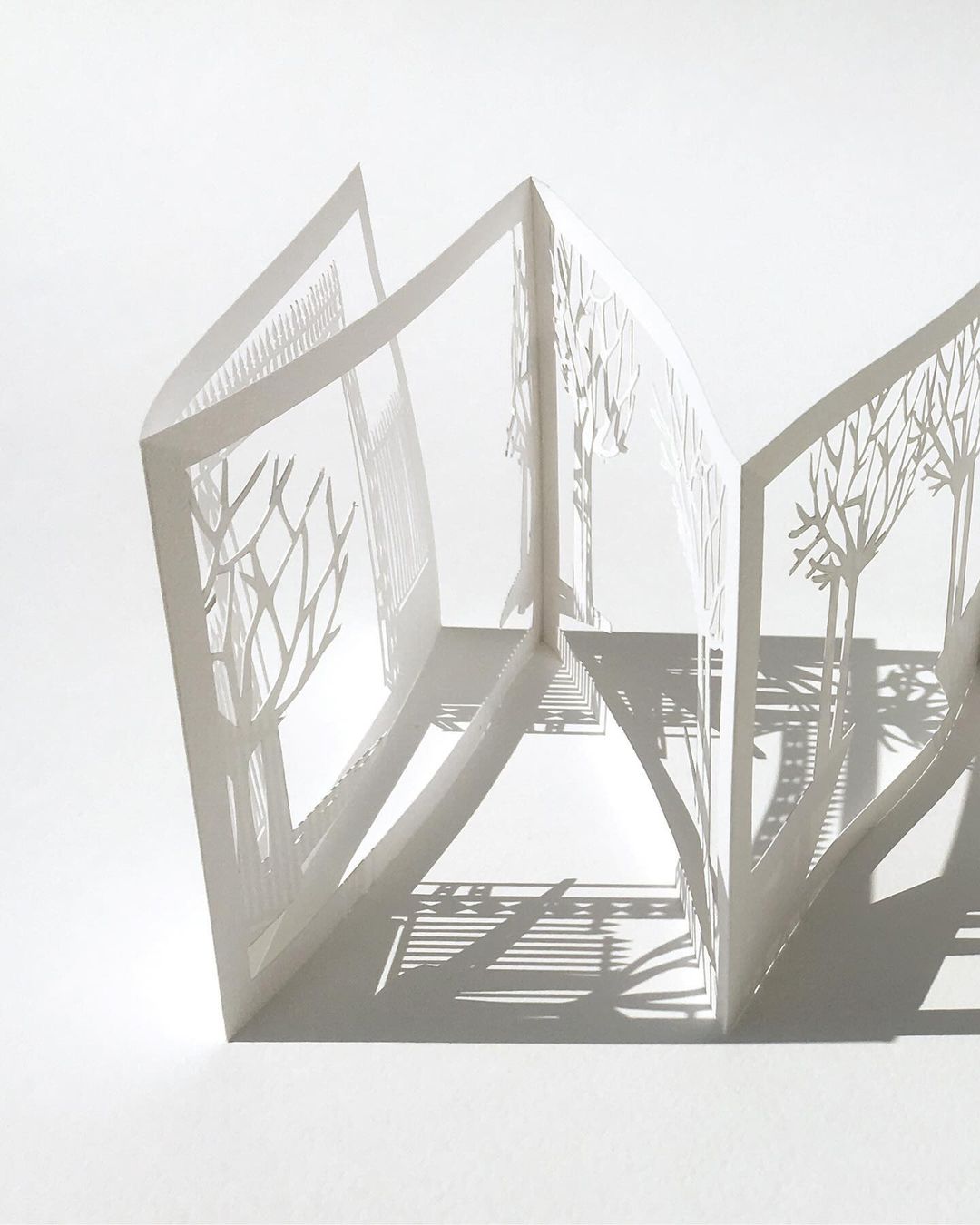 Paper art featuring cut-out trees arranged in zigzag formation, creating a miniature paper landscape with shadows on a white backdrop.