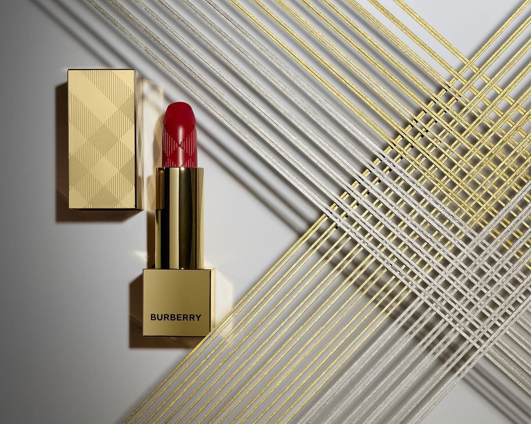 Burberry lipstick in gold packaging with a red product stands against a white backdrop with a gold and silver woven design.