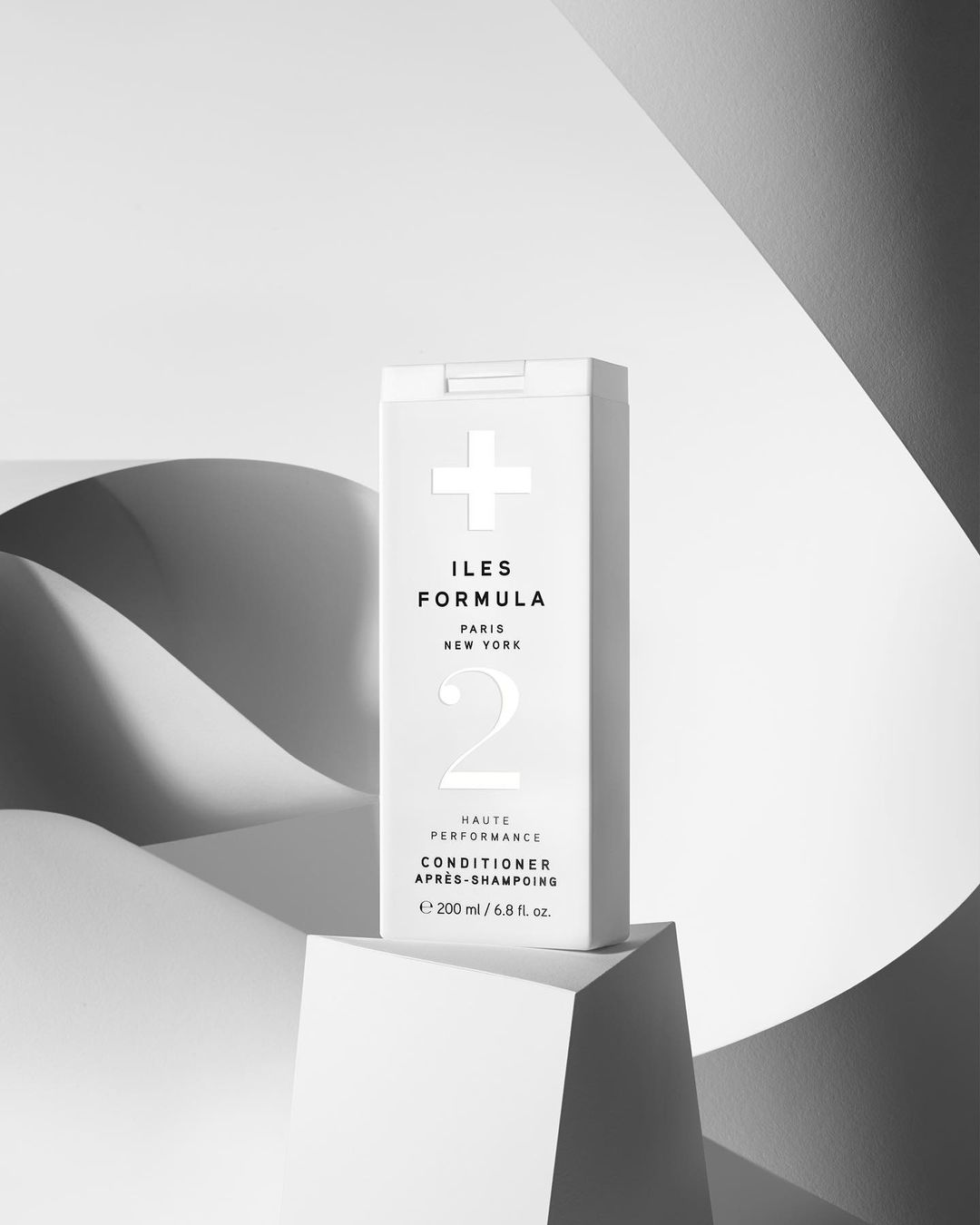 Iles Formula conditioner product shot in a minimalist setting. White bottle on a geometric block, neutral color palette.