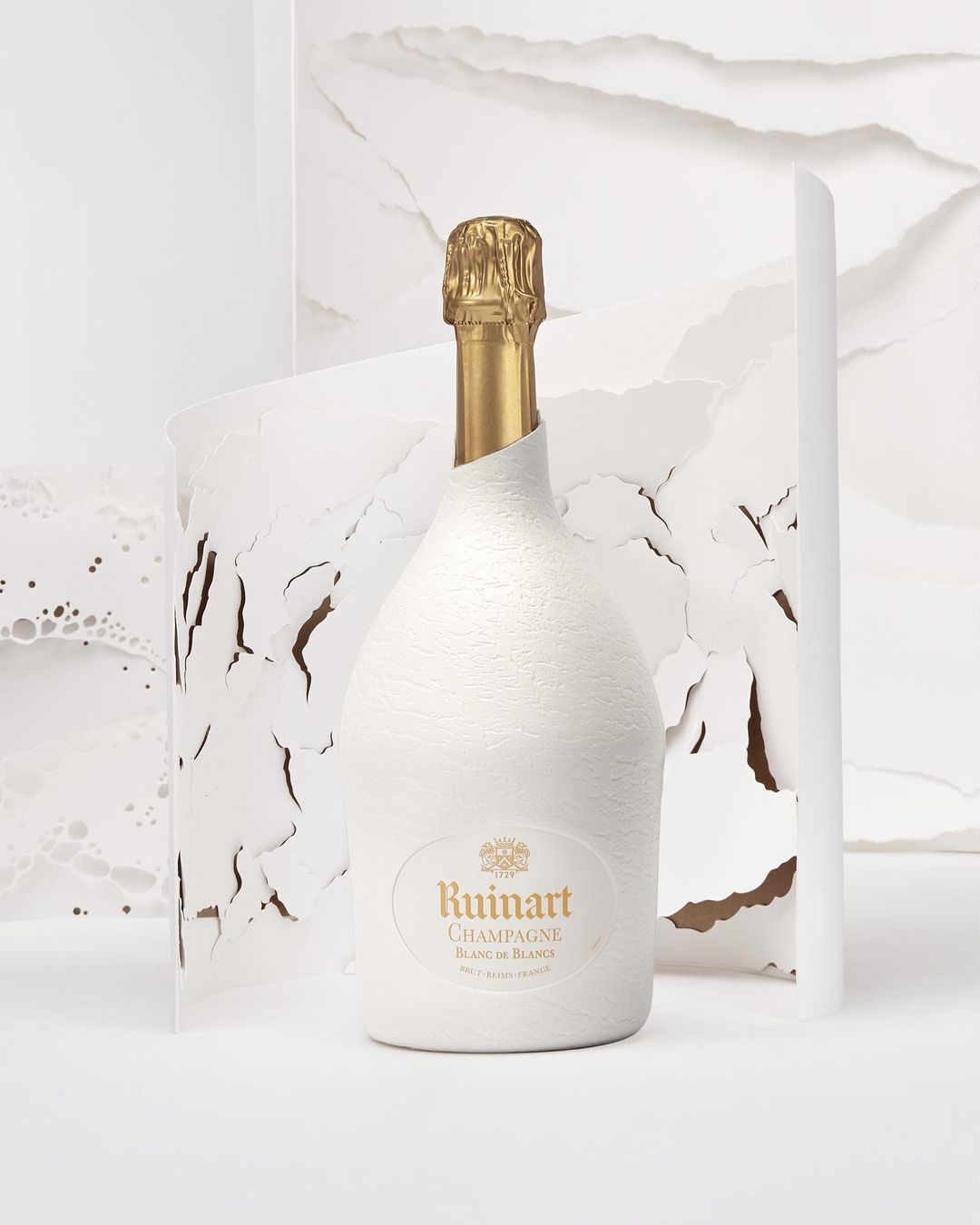 Studio shot of a Ruinart champagne bottle encased in textured white material with a gold foil top against a torn paper background.