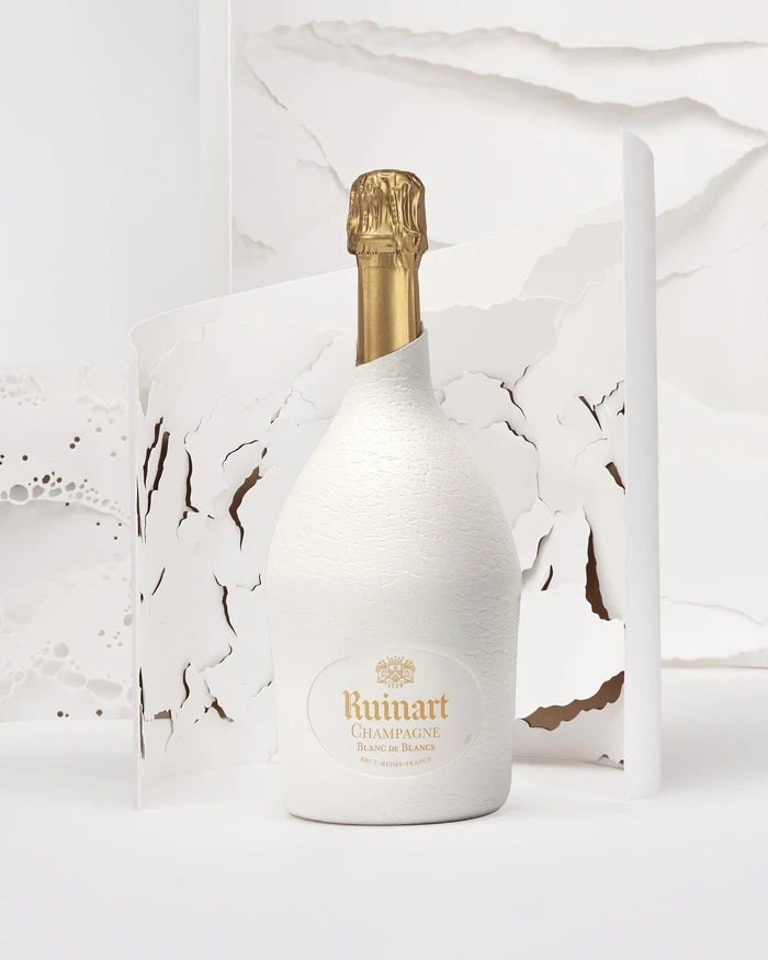 White textured champagne bottle with gold foil neck in front of layered torn paper sculpture by Cloé Vriet