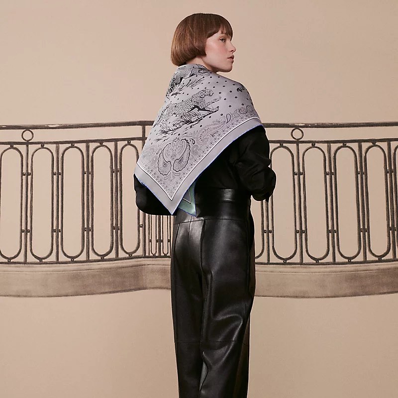 Fashion shot of a woman wearing a grey patterned scarf, a black top, and black leather pants, standing in front of a beige background.
