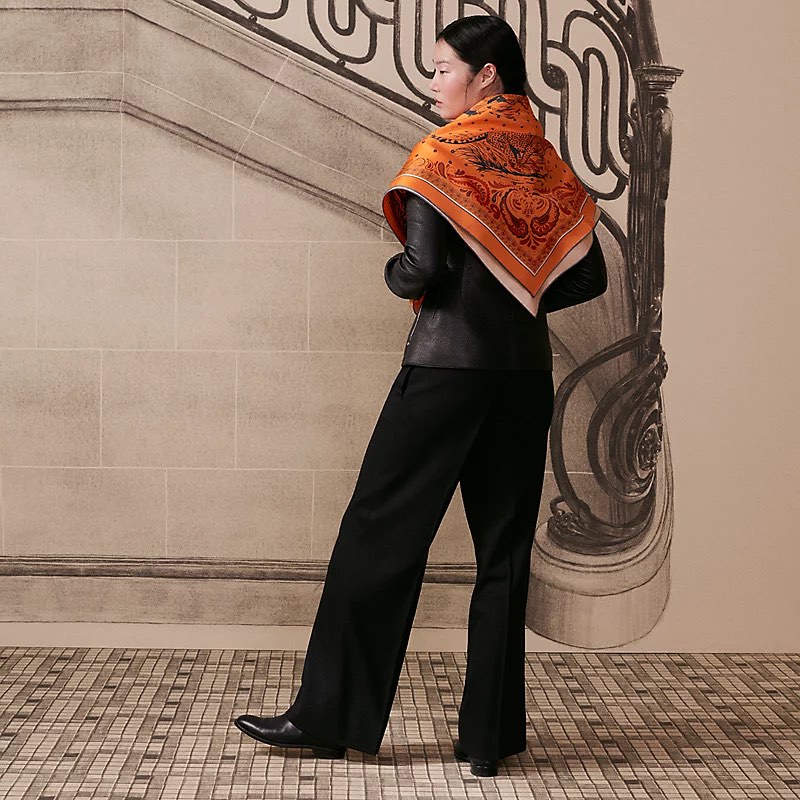 Fashion shot of an Asian woman wearing a black jacket and pants, accessorized with an orange printed silk scarf, indoors.