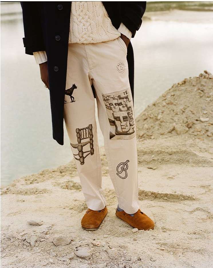 Person in light beige trousers with graphic prints, a cream-colored sweater, and a dark coat, standing on a sandy surface outdoors.