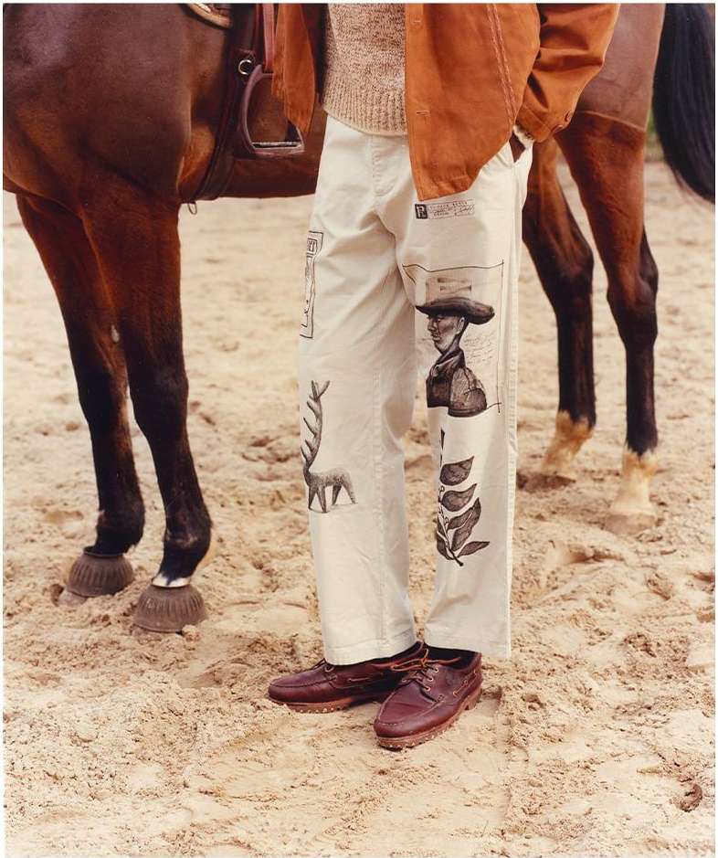 Man wearing illustrated white pants and brown leather shoes standing next to a horse on a sandy ground