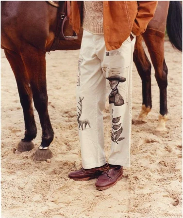 Man wearing illustrated white pants and brown leather shoes standing next to a horse on a sandy ground