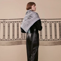 Woman wearing a patterned silk scarf and black leather trousers standing before an ornate metal balcony railing