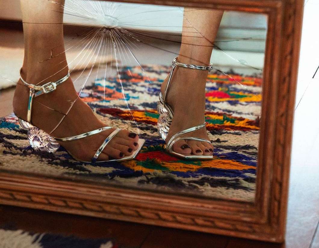 Reflection in broken mirror of adult woman wearing silver ankle strap high heel sandals on a colorful shag rug indoors.