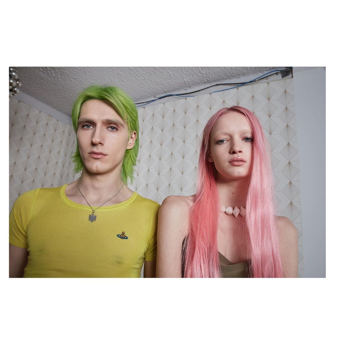 A couple poses in a photo studio with vibrant green and pink hair. The man wears a yellow shirt, and the woman has on a light-colored beaded necklace.