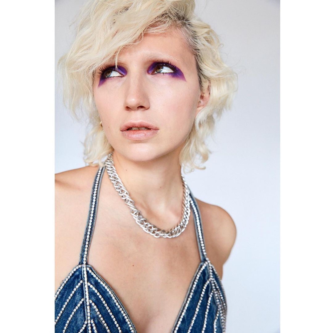 Close-up of a young adult woman with platinum blonde hair and purple eye makeup, wearing a denim top and chain necklace.