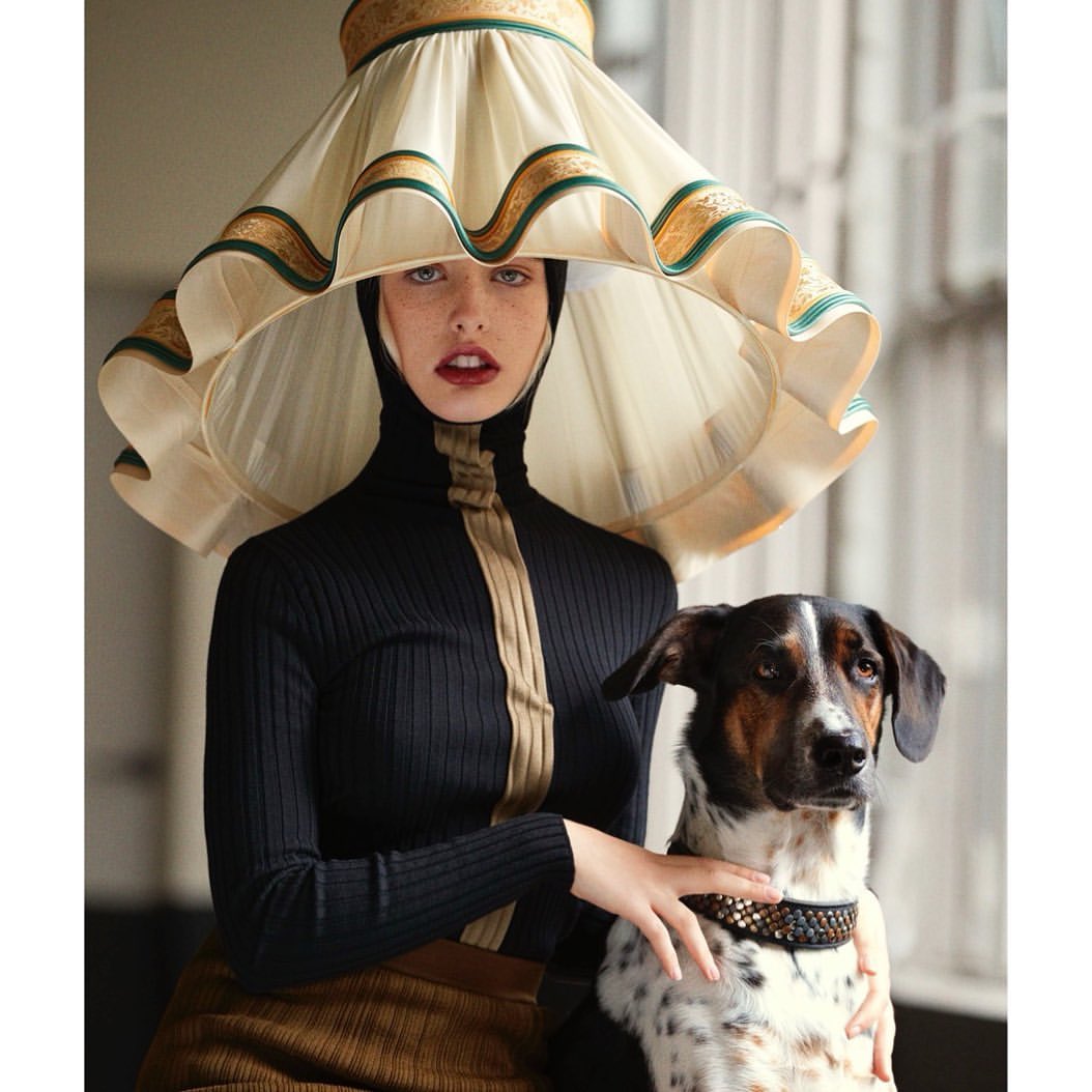 Woman in black turtleneck and tan skirt poses with her dog in front of a window. She wears a lampshade as a hat.