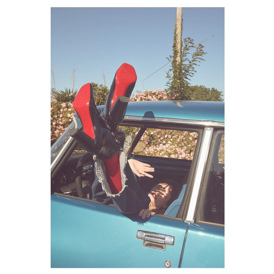Fashion shot of a woman partially visible hanging upside down out of a blue car window wearing black pants and red-bottom stilettos.
